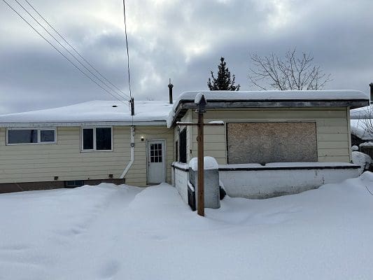 Here's an alt tag for the image: Snow-covered house, boarded-up window.