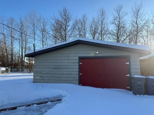 Snow-covered garage with red doors, trees.
