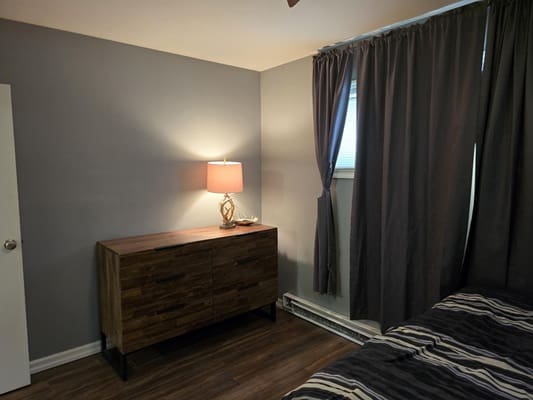 Bedroom with dresser and lamp, dark curtains.