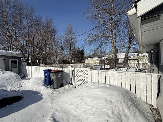 Snowy backyard with trees and trash bins.